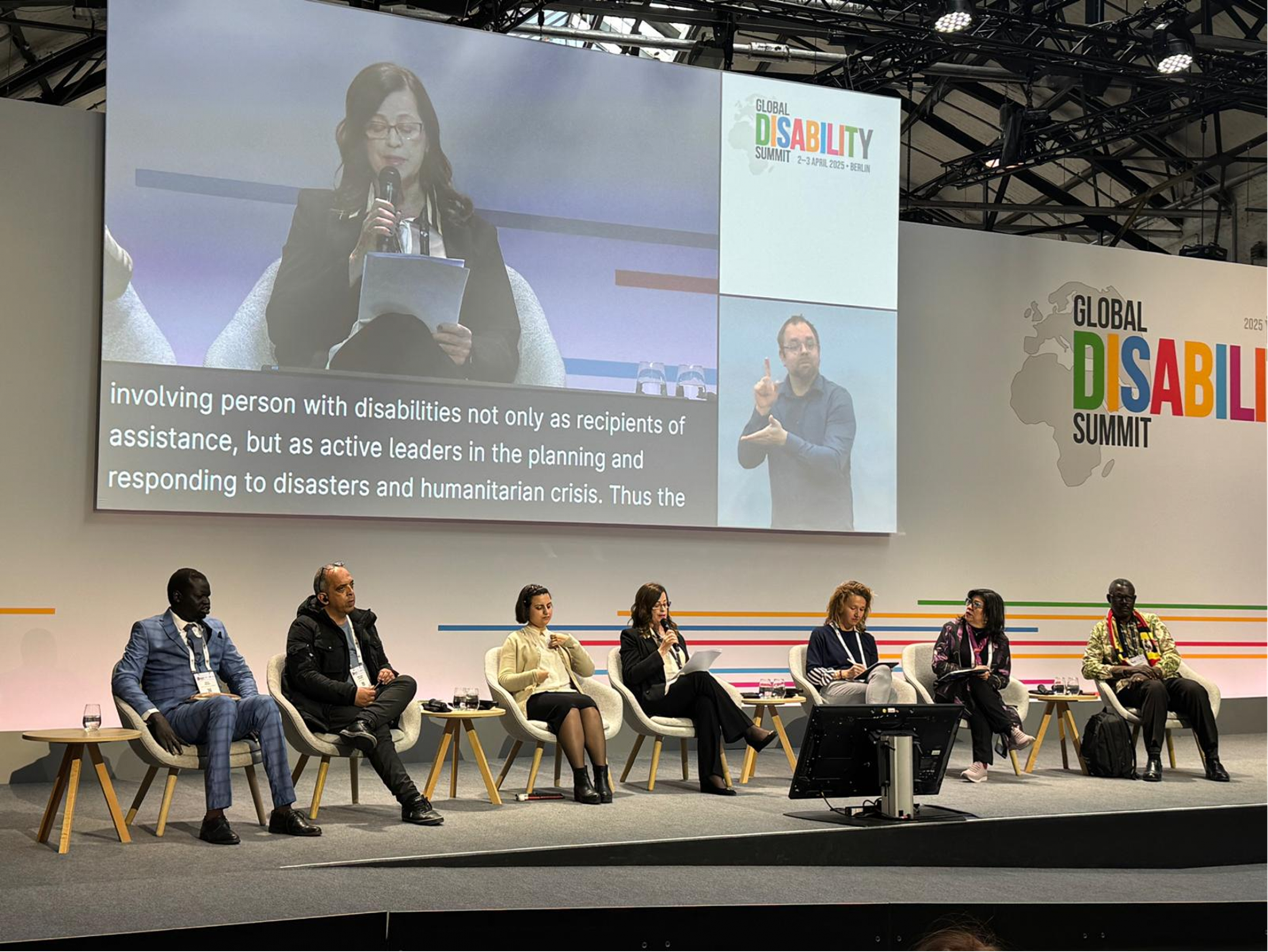 The illustration takes place in a refugee camp. There is a registration booth where a woman wearing a headscarf is sitting at a desk taking information from a woman in a wheelchair also wearing a headscarf and queuing. Behind her there is a man and a woman standing, a woman with two children talking in front of an accessible bathroom. In front of them there is an accessibility ramp and three people: A man providing information with a megaphone while a woman translates this information into sign language for a man with hearing impairment. There is a tent where the registration booth is located and also a waiting area where two men and a woman with visual impairment are seated. A truck carrying food supplies enters the refugee camp and it can be seen that they are unloading several packages of food.    