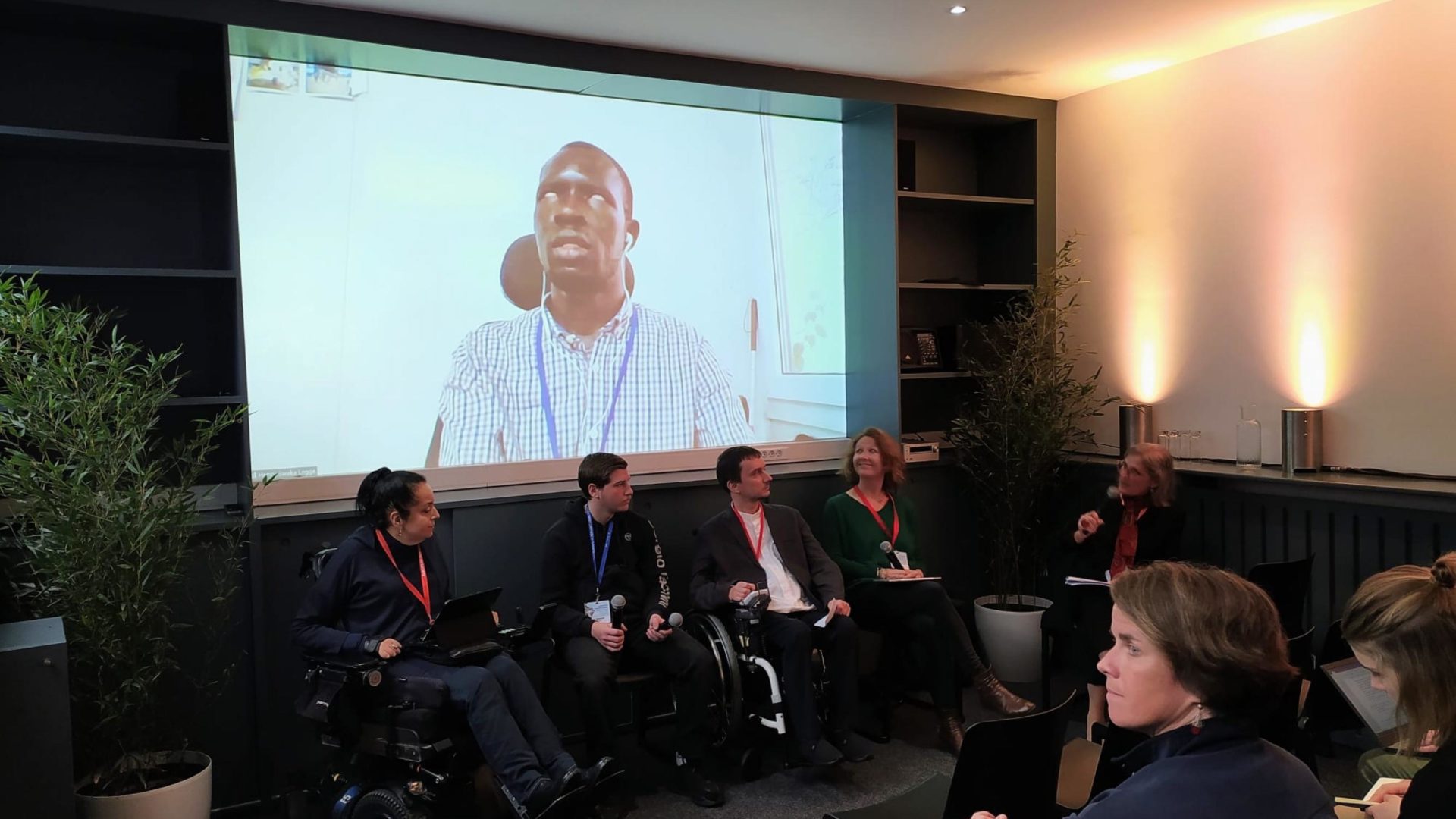 Picture of the round table at the European Humanitarian Forum. Three panelists, one assistant and one moderator are present in the room in Brussels, one remote panelist is displayed on a large screen. Two of the panelists in the room are wheelchair users.