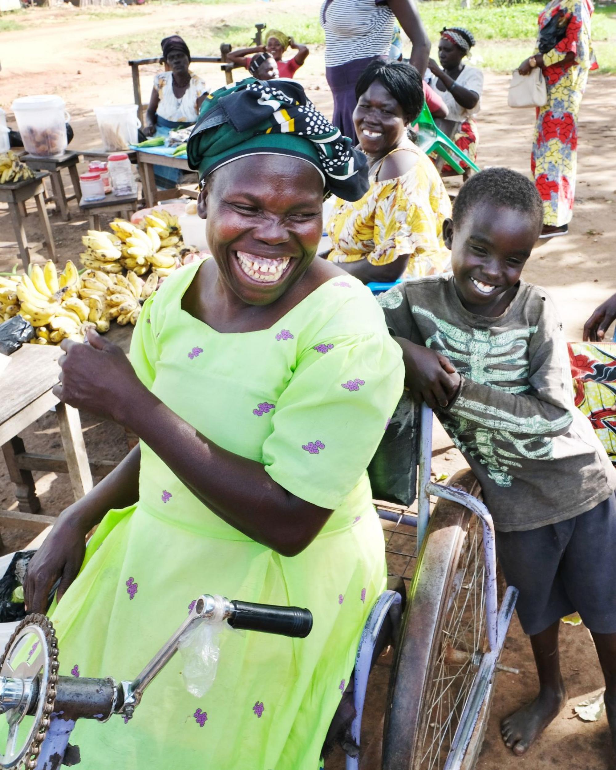 Uganda: Foto einer älteren Frau, die auf einem Holzstuhl sitzt. Sie hält eine Krücke in ihrer Hand. Sie hat eine Seh- und physische Beeinträchtigung. Neben ihr sitzen ein junger Mann und eine junge Frau von Handicap International. Ein weiterer junger Mann schaut in die Richtung der älteren Frau. Er ist kein Mitarbeiter der Organisation.