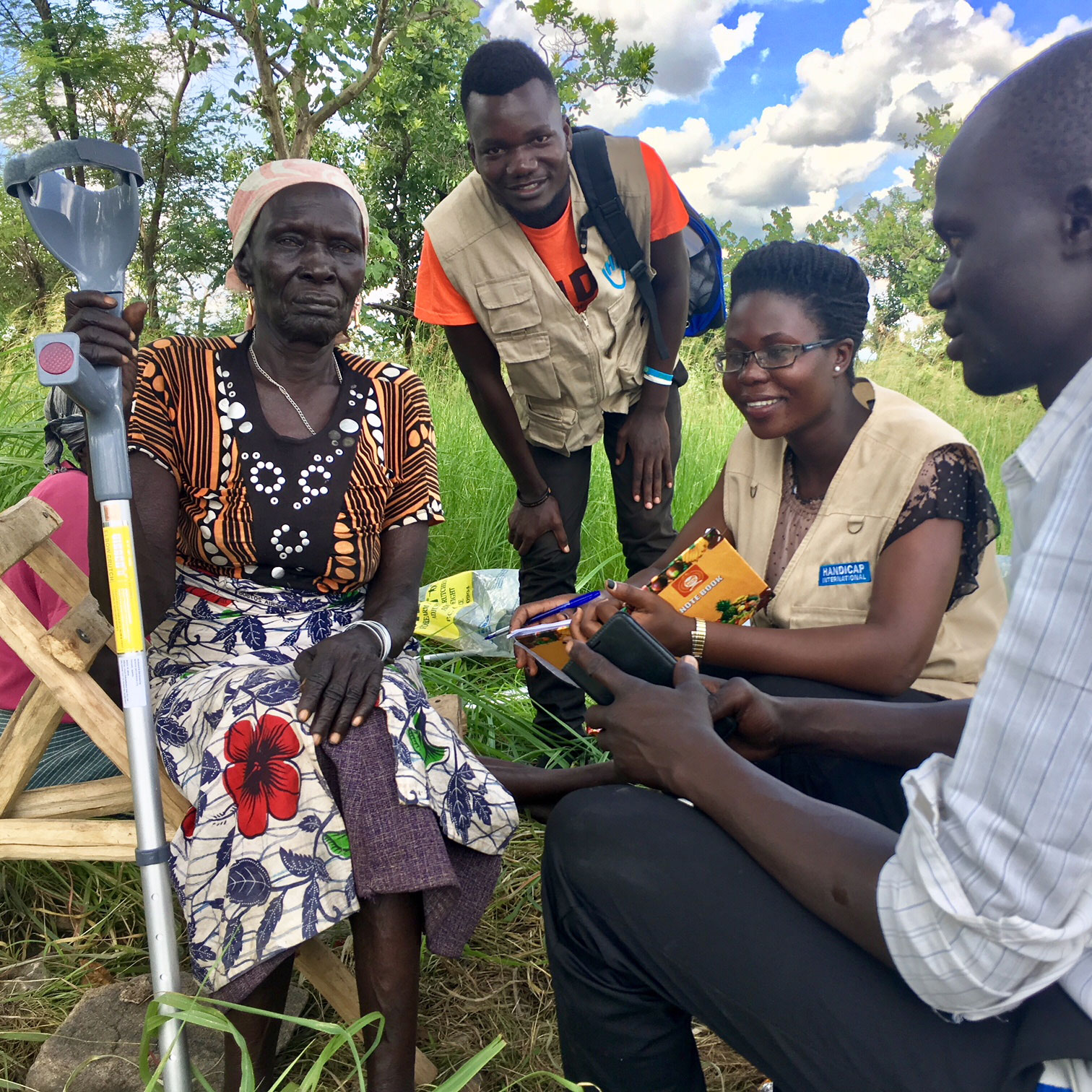Uganda: Photo of an older woman sitting on a wooden chair. She holds a crutch in her hand. She has visual and physical impairments. A young man and a young woman from Handicap International sit next to her. Another young man looks in the direction of the older woman. He is not an employee of the organization.